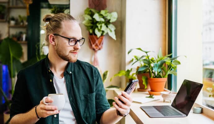 Hombre que bebe una taza de café mientras utiliza un teléfono móvil protegido con Norton 360 for Mobile